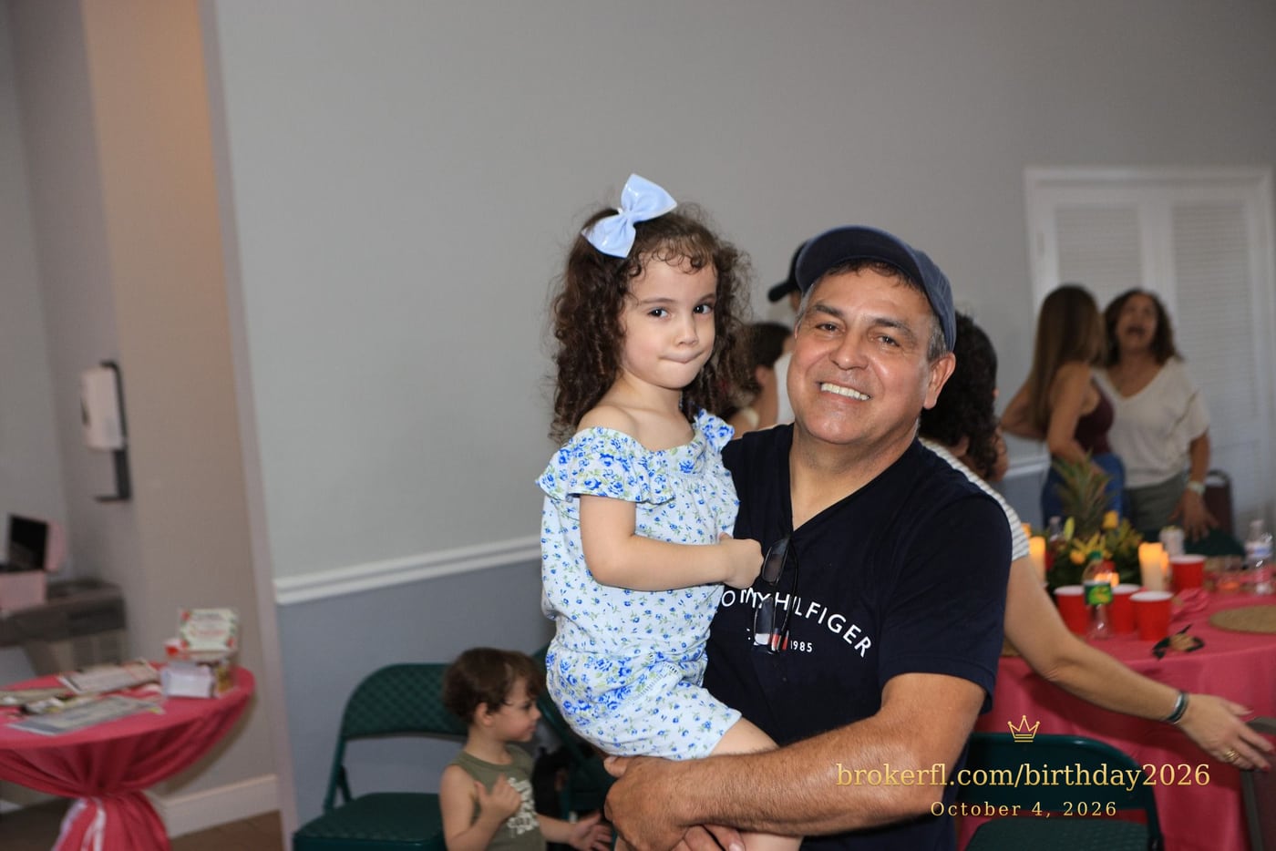 Adult holding a young guest in a blue dress at the celebration — Karina Leal's birthday, October 4, 2026.