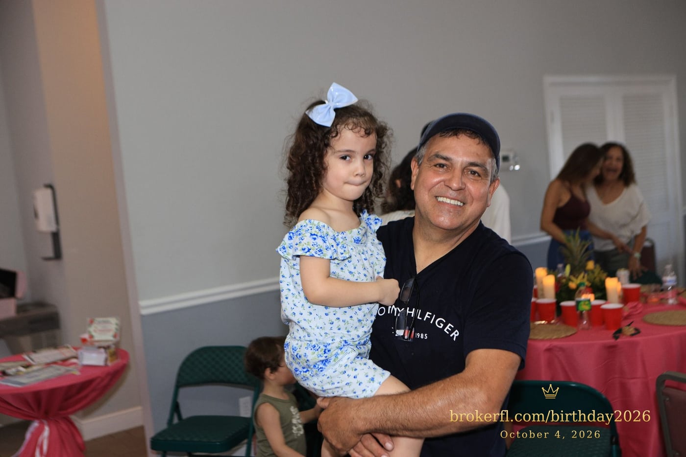 Adult holding a young guest in a blue dress at the celebration — Karina Leal's birthday, October 4, 2026.